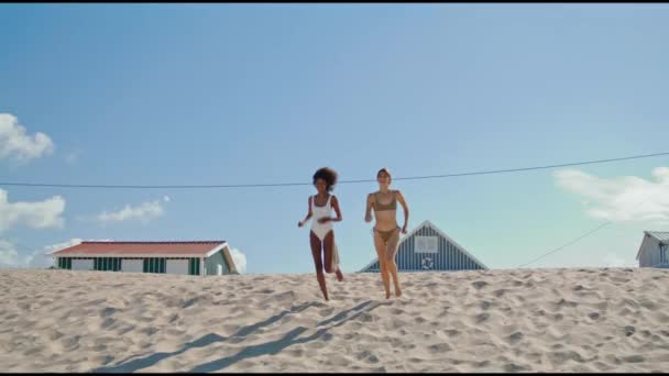 ビキニでビーチを走ってるふたり 幸せな女の子は海の海岸で夏を過ごす スリム多民族の友人のパートナーは 海岸で週末を楽しんで泳ぎに行く 一緒に楽しみを持っているケアフリーガールフレンド — ストック動画