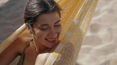 Woman chilling hammock beach alone portrait. Smiling beautiful girl closing eyes relaxing in bikini summer sunlight. Serene attractive lady dreaming looking camera at seashore. Seaside lounge concept.