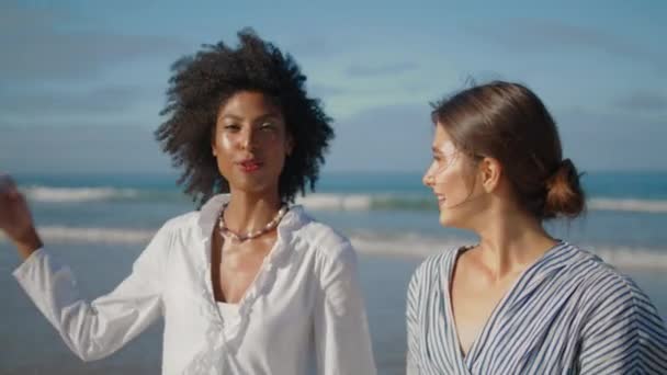 两个女孩在谈论近岸特写 无忧无虑的女同性恋夫妇在阳光下在海滨散步 微笑的多种族女性伴侣分享去沙滩的新闻 最要好的朋友在户外度周末 — 图库视频影像