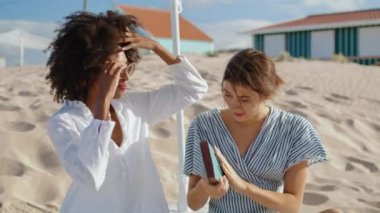 Two women holding book resting beach picnic. Beautiful multiethnic lgbt couple talking discussing novel in summer sunlight. Carefree stylish girls enjoying ocean vacation. Weekend leisure concept.