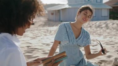 Friends spending beach time discussing book. Closeup smiling attractive girl taking off sunglasses talking with african american partner at sandy ocean coast. Lgbt couple rest on sunny summer weekend