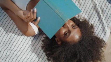 Plaj kızı kitap okuyor, battaniyenin üstünde yatıyor. Neşeli, güler yüzlü kadın güneş ışığının altında yüzünü gizliyor. Çekici Afro-Amerikan poz verme romanı. Tatil hobisi kavramı.