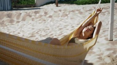 Relaxed girl sunbathing hammock on sandy beach. Beautiful carefree woman resting looking camera on summer vacation. Happy dreaming tourist lying enjoying sunlight. Weekend leisure at seashore concept.