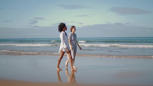 Lgbtカップルは日光の中で海の海岸に行く 無料のガールフレンドは 夏の週末に砂浜を歩く会話をお楽しみください 海の岸でニュースを共有する多民族の女性を笑顔 女の子の友情の概念 — ストック動画