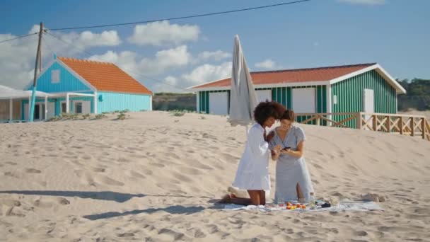 同性恋夫妇使用智能手机检查海滩图片 两个女孩在海滨休息 周末在沙滩上野餐 快乐的多种族伴侣检查社交媒体的帖子 快乐的闲暇时间 — 图库视频影像