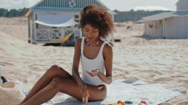 Kumsalda güneş kremi süren güzel bir kız. Şık bir kadın okyanus kıyısında tek başına güneşleniyor. Afro-Amerikan masaj yapan rahat bir vücut ve UV koruma kremi. Tatil gevşeme kavramı