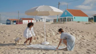 Girlfriends spread picnic blanket on sandy beach. Two girls enjoying summer sunny day relaxing at seashore together. Romantic multiethnic love couple talking resting outdoors. Idyllic weekend morning.