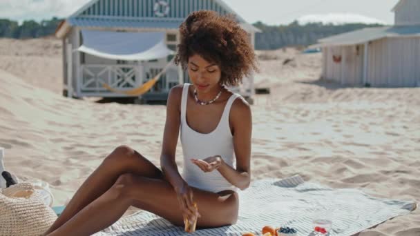 ビーチで日焼け止めを塗る美しい女の子 海の岸で一人で日光浴スタイリッシュな女性 休暇中にUv保護クリームとリラックスしたアフリカ系アメリカ人のマッサージボディ 休日のリラクゼーションコンセプト — ストック動画