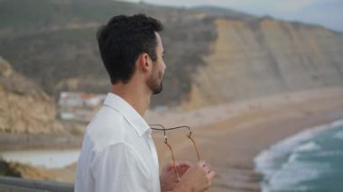Calm tourist taking sunglasses at ocean beach closeup. Sexy guy looking summer nature. Bearded man enjoying sea shore travelling alone. Peaceful relaxed model resting marine landscape view vacation