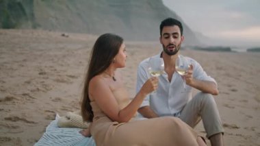 Romantic couple celebrating vacation at seashore. Positive family holding wineglasses drinking alcohol at sandy beach picnic. Handsome man telling jokes to laughing woman. Spouses spending anniversary