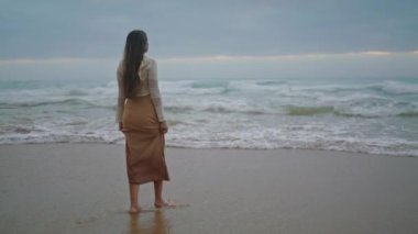 Romantic girl looking ocean view at evening. Dreamy woman walking on stormy beach wide shot. Female model spending vacation at seaside alone. Brunette lady enjoying of scenery marine landscape