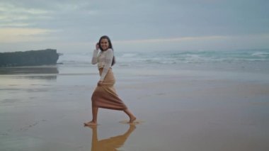 Positive girl laughing beach wide shot. Carefree woman raising hands at cloudy marine view place. Cheerful model having fun at evening seashore. Relaxed lady enjoying freedom on ocean nature walk