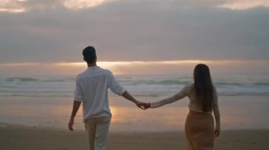 Young spouses relaxing sunbeams beach at cloudy evening. Happy lovers holding hands walking together at sea coast back view. Loving woman man romantic honeymoon date at ocean side. Love relationship 