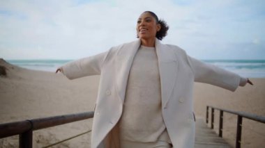 Happy woman spinning open arms on ocean beach. Smiling cheerful model walking pathway enjoying autumn seaside trip alone. Beautiful confident african american rejoicing celebrating life at calm shore.