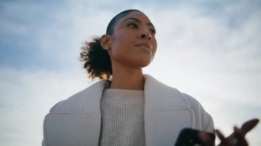 Worried woman checking phone waiting for reply closeup. Black hair model looking camera bottom view. Pensive girl in sunlight holding smartphone making decision. African american send email message 