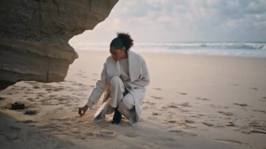 Thoughtful woman drawing sand at ocean cliff. Black hair traveler resting beach enjoying morning walk. Focused african american dreaming meditate outdoors. Lonely beautiful girl relax spend time alone