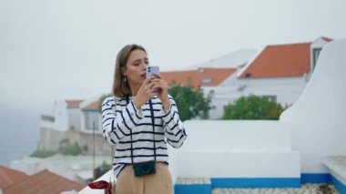 Gezgin kız beyaz çatıda akıllı telefon fotoğrafları çekiyor. Kasabayı gezen turistler deniz köyü manzarasında tatil fotoğrafları çekiyor. Odaklanmış blog öğrencisi sosyal medya içeriği oluşturur. Tatil eğlencesi konsepti