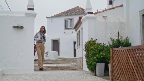 游客女孩漫步在古老的冥想城大街上 摄影师在漂亮的白色房子里检查相机 漂亮的女人正在游览风景秀丽的小镇 享受暑假的旅客 — 图库视频影像