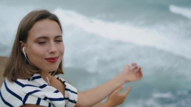 Gülümseyen milenyum müziği okyanus kıyısında yakın çekim. Mutlu kız, hafta sonu tatilinde deniz uçurumunun kenarında sakin bir şekilde kameraya bakar. Romantik genç kadın deniz kıyısının tadını çıkarıyor.