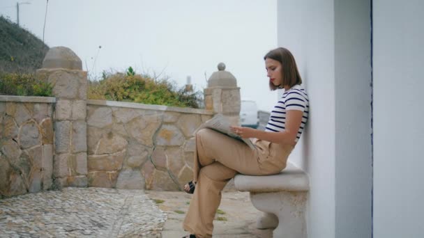 女人在阳台上看晨报 一个松懈的女孩在户外喝咖啡 欣赏春天周末平静的气氛 美丽的嬉皮士游客在暑假时持着当地杂志休息 — 图库视频影像