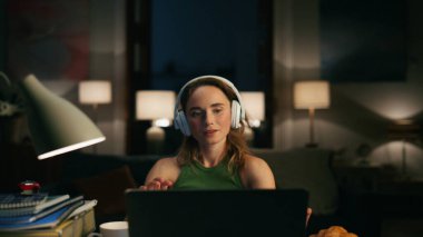 Genç bir uzman gece yakın planda dizüstü bilgisayarda çalışıyor. Modern bayan yaratıcı akşam ortamında bilgisayar izliyor. Podcast dinleyen pozitif kulaklıklı kız serbest çalışmanın tadını çıkarıyor.