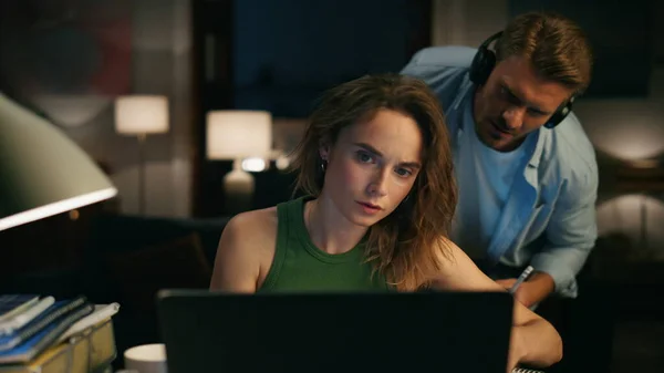 Freelance lady writing text at evening flat closeup. Headphones man distracting from work peeping screen. Husband making noise asking attention. Thoughtful woman browsing laptop ignoring at home