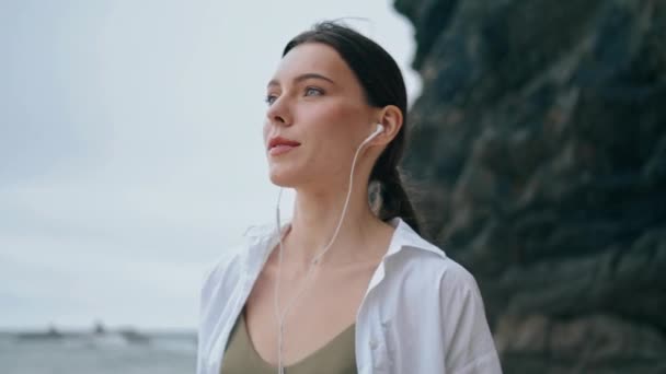 快乐无忧无虑的女人带着耳机在岸边散步 享受着美妙的音乐特写 迷人的小女孩穿着白衬衫 用现代耳机听着歌曲的画像 漂亮的女士带着耳机 — 图库视频影像
