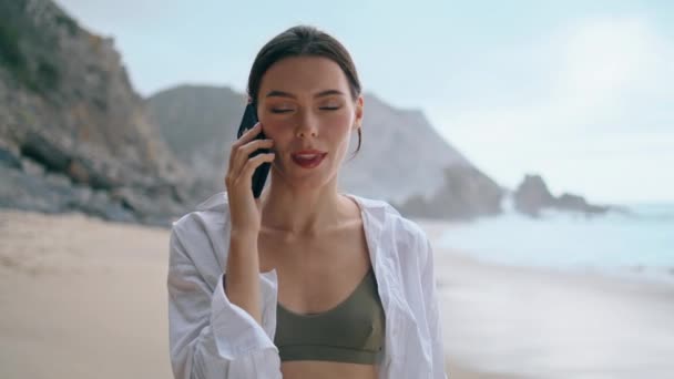 积极迷人的女孩在阴暗的海滨用智能手机与微笑近距离交谈 年轻镇定的女人站在沙滩上打电话 漂亮的女士喜欢在岸上的移动对话 — 图库视频影像