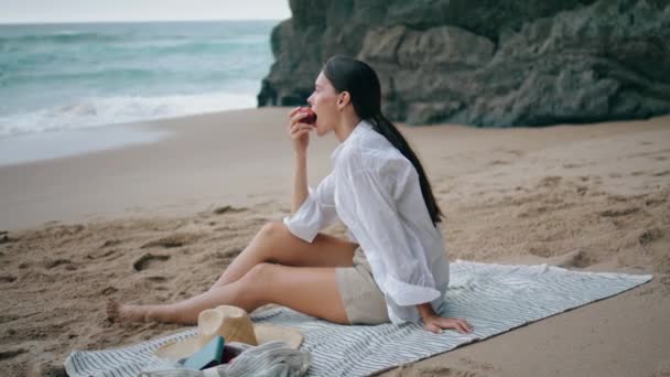 在海滩野餐时 坐在阴天的毛毯下吃苹果的年轻姑娘松了口气 迷人而宁静的女人在沙滩上欣赏着海浪带来的新鲜水果 沉着的女士一个人在海滩上放松一下 — 图库视频影像