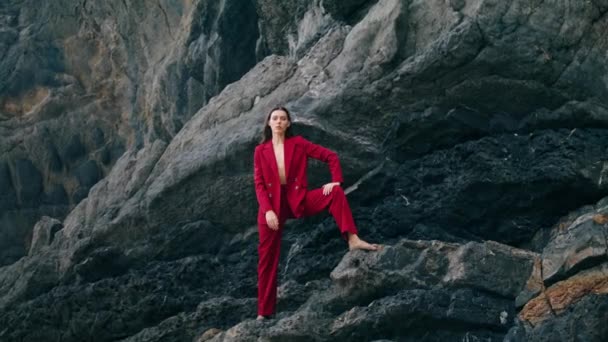 エレガントなファッションの女性の崖の石の棚悲観的な灰色の週末に足を置くに立っている 風光明媚な岩のポーズで赤いスーツの魅力的なスタイリッシュな女の子 自然の中で自由さを感じる美しいモデル — ストック動画