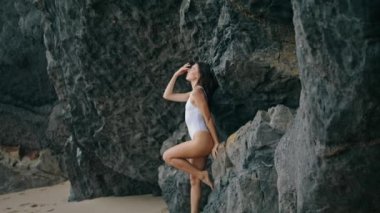 Taş kayaya yaslanmış sahilde poz veren seksi genç kadın. Deniz kıyısında beyaz mayo giyen çekici, şehvetli bir model. Uçurumun kenarındaki kumsalda duran muhteşem seksi kız. Güzellik konsepti.