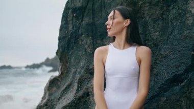 Güzel deniz manzarası seyreden şehvetli genç bir kadın kayaya yakın duruyor. Bulutlu sahilde duran beyaz seksi mayolu çekici nazik kız. Muhteşem genç model fırtınalı okyanus dalgalarını izliyor..