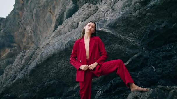 悲観的な性質で足を岩の縁に置く豪華なスタイリッシュなモデル 裸体でエレガントな赤いスーツを着た官能的な女性が自信を持ってカメラを見ている 魅力的なファッション女の子立って崖 — ストック動画