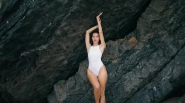 Deniz kıyısında poz veren güzel, şehvetli bir model yüksek kayalara el kaldırıyor. Taş deniz kıyısında duran beyaz mayo giyen çekici seksi kadın. Muhteşem ciddi kız rahatlatıcı yaz tatilinin tadını çıkar..