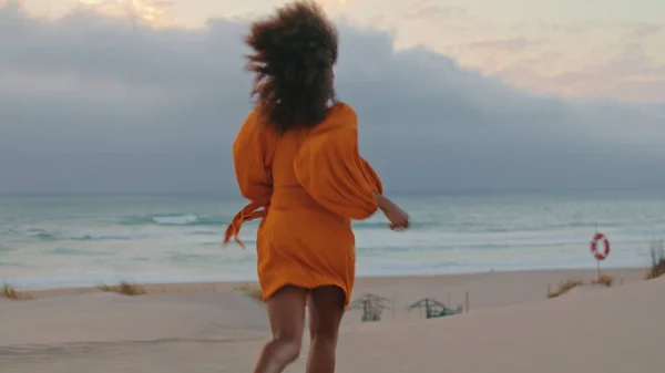 快乐的女人在灰蒙蒙的天空中逃离沙滩 穿着橙色连衣裙在海滨跑步的快乐的非洲裔美国女孩 在夏夜嬉笑着 卷曲的黑发在大自然中享乐 — 图库照片