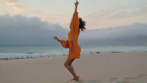 Emotional girl contemporary dancer waving hands performing on sand seashore summer evening. Smiling flexible african american woman wearing orange dress dancing modern style on gloomy beach.