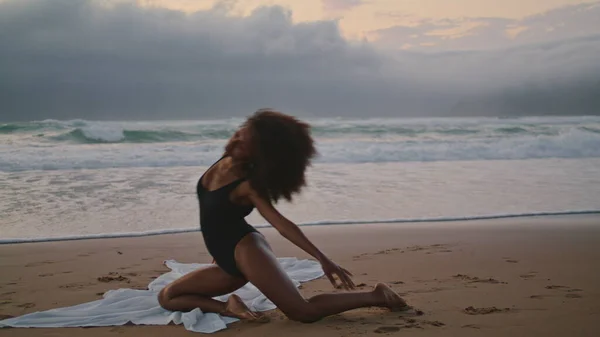 性感的非洲裔美国女孩在沙滩上跳舞 夏日阴郁的夜晚 卷曲柔韧的黑发 在激情表演中创造杂技元素 身穿黑色泳衣的瘦女人优雅地移动着身体 — 图库照片
