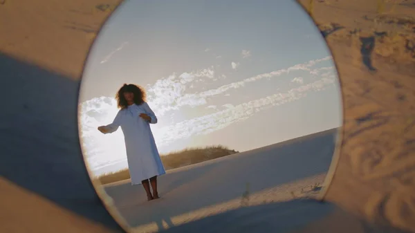 Round mirror reflecting woman dance at sand desert in front beautiful sunset. African american girl wearing white dress making graceful hands movements standing in wilderness. Performance on nature.