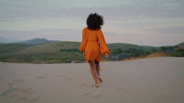 在夏夜黄昏时穿着橙色连衣裙在沙漠上慢跑的女子的背影 未知的卷曲女孩蹲伏在沙滩上 在漆黑的夜晚 活跃的非洲裔美国黑人在美丽的沙丘上放松一下 — 图库照片