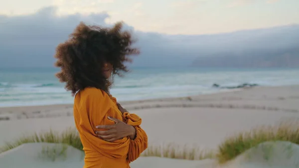 stock image Attractive relaxed model woman posing wearing orange dress at overcast nature close up. Happy african american girl smiling on camera touching lush curly hairstyle. Brunette standing on sand seashore.