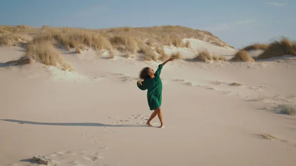 Young sensual woman performing emotional dance in hot desert alone. Sporty african american girl moving mesmerizingly wearing black dress. Curly pretty brunette dancer spinning body on sand dunes.