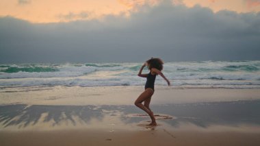 Graceful african american girl dancing on wet sand near ocean waves summer cloudy evening. Curly attractive brunette in black swimsuit performing sensual dance on gloomy seashore. Modern performance.