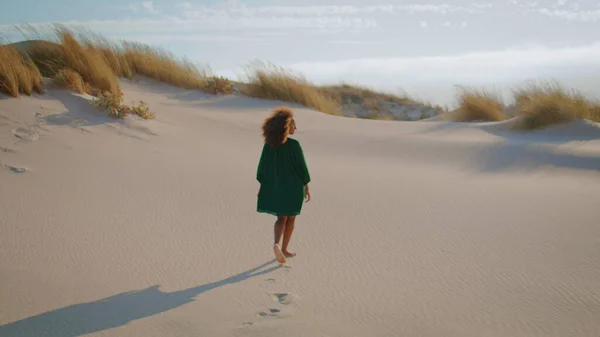 Back view african american woman walking desert dunes in black dress. Unknown young girl stepping on sand making footmarks summer windy day. Relaxed curly brunette enjoy beautiful nature in wilderness