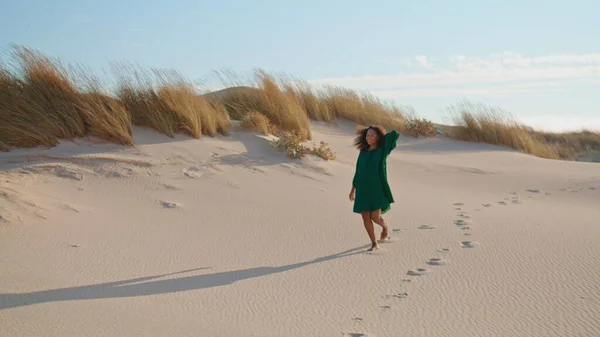 African american girl jogging down sand hill leaving footprints at summer desert. Attractive relaxed woman walking on dunes wearing black dress. Young brunette enjoy holiday at wilderness nature.