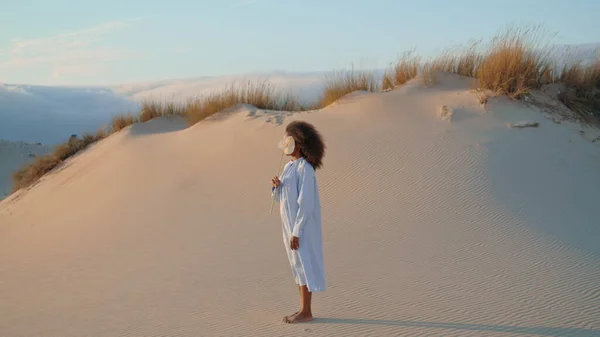 African american actor woman posing on sand desert with white flower summer windy day. Calm girl standing at dunes with dry grass holding beautiful calla. Young brunette enjoy nature looking distance