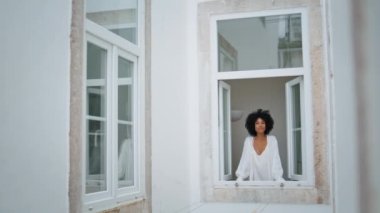 Pozitif kadın pencereye bakıyor. Kıvırcık gülümseyen kadın dışarıda dikizlemekten hoşlanıyor. Mutlu Afro-Amerikan modelleri sabah şehir manzarasının rahatlamasını seyrediyor. Günlük rutin kavram