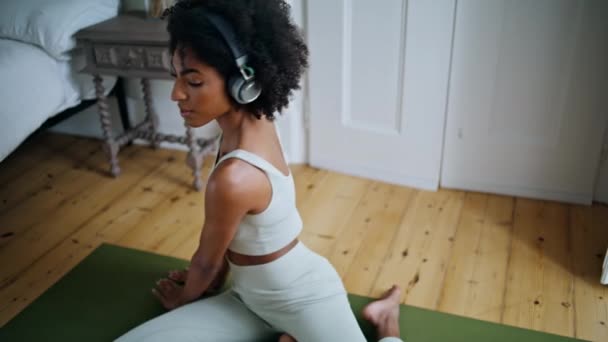 年轻的女士在室内的垫子上伸展身体 黑头发女人坐在橡胶地毯上做瑜伽练习 耳机女孩早上一个人听歌曲训练 灵活的模型热身肌肉 — 图库视频影像