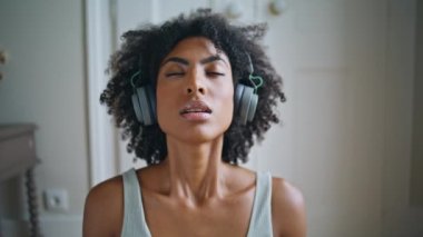 Serene modeli yoga solunumu yapıyor. Afrikalı bir kadın tek başına meditasyon yaparken beyaz odada yavaşça nefes alırken görülüyor. Kapalı gözlü kız derin nefes alıp veriyor kulaklıkları dinliyor rahat müzik dinliyor. 