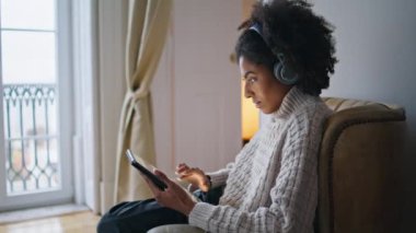 Rahat evdeki odaklanmış kız kaydırma tahtası. Serbest çalışan, tablet seyreden, sakince tek başına çalışan bir kadın. Afro-Amerikan kulaklıklı bayan rahat koltukta oturmuş müzik dinliyor. Ev içi rutin kavram