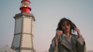 Beautiful girl resting lighthouse view. Casual young woman looking camera adjust curly hair. Worried pensive female contemplating walking seaside. Serious traveler turning enjoying evening trip alone